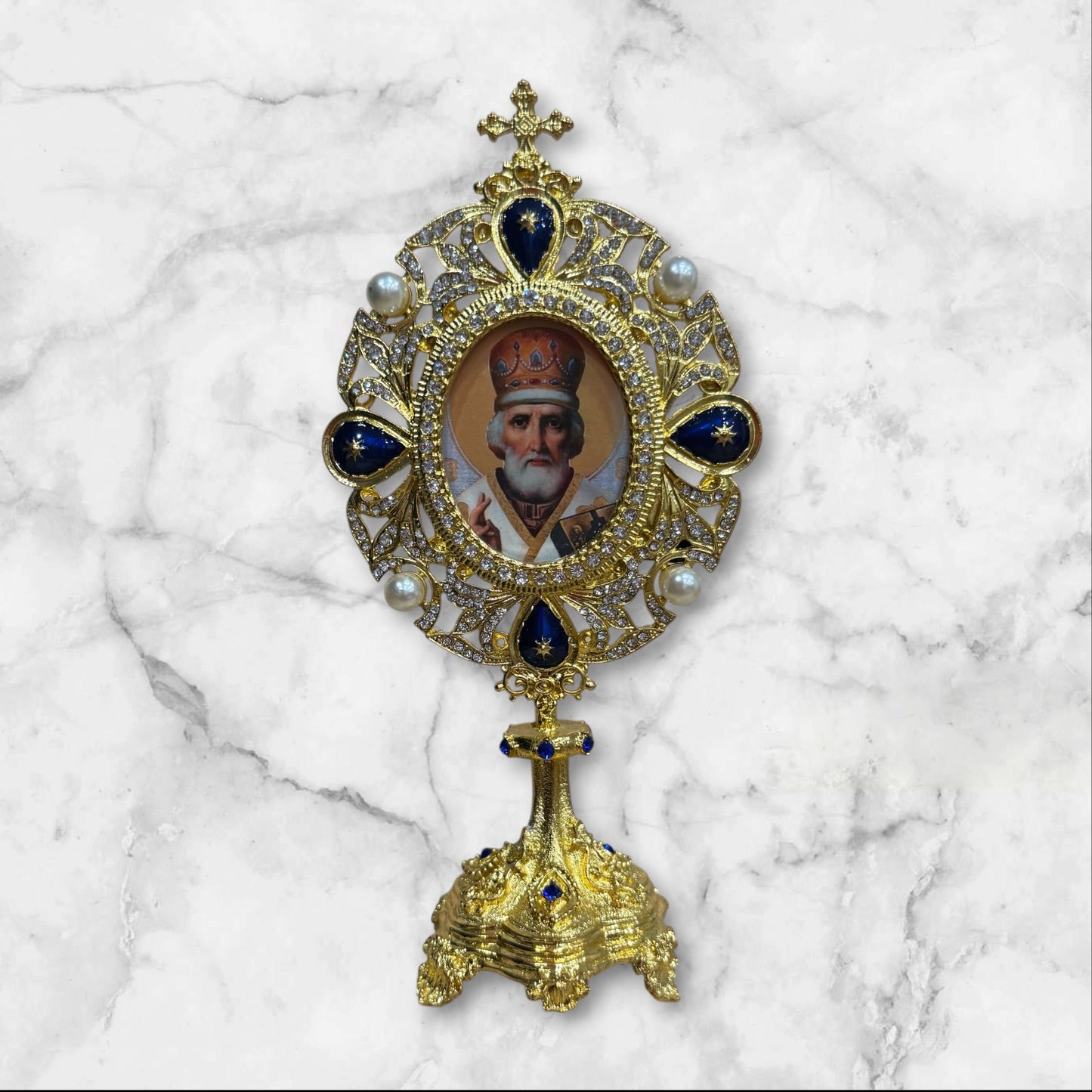 Jeweled oval standing icon of Saint Nicholas featuring an ornate gold-tone frame with blue enamel accents, pearl details, rhinestones, and a decorative freestanding base.
