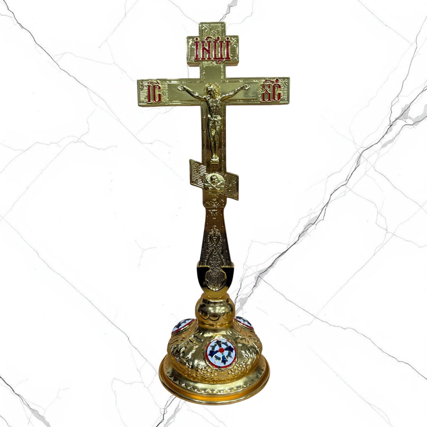 An ornate gold-toned Orthodox Christian cross with a crucifixion figure and red and blue enamel details, set against a white marble-textured background.
