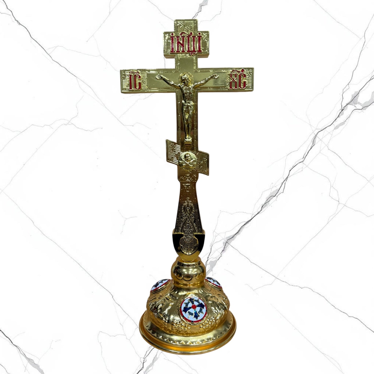 An ornate gold-toned Orthodox Christian cross with a crucifixion figure and red and blue enamel details, set against a white marble-textured background.